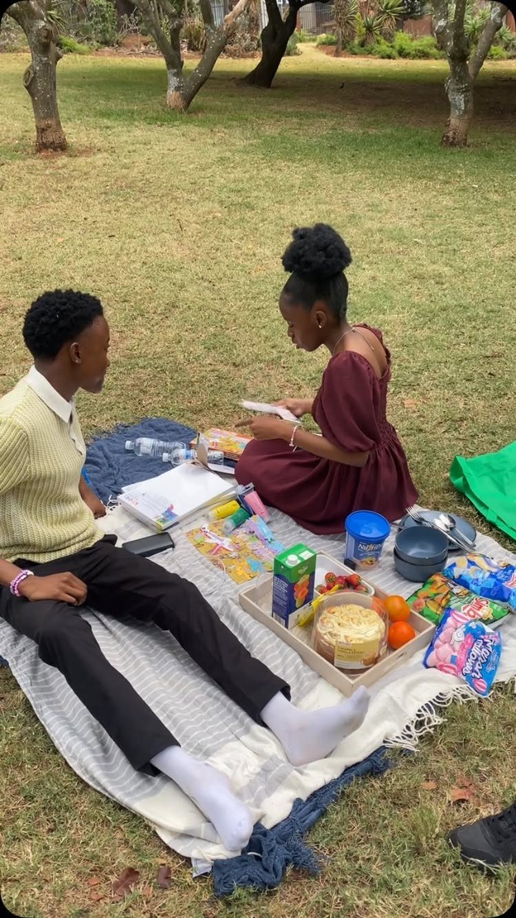 Couple sitting on a blanket in a park with a picnic spread of food.
