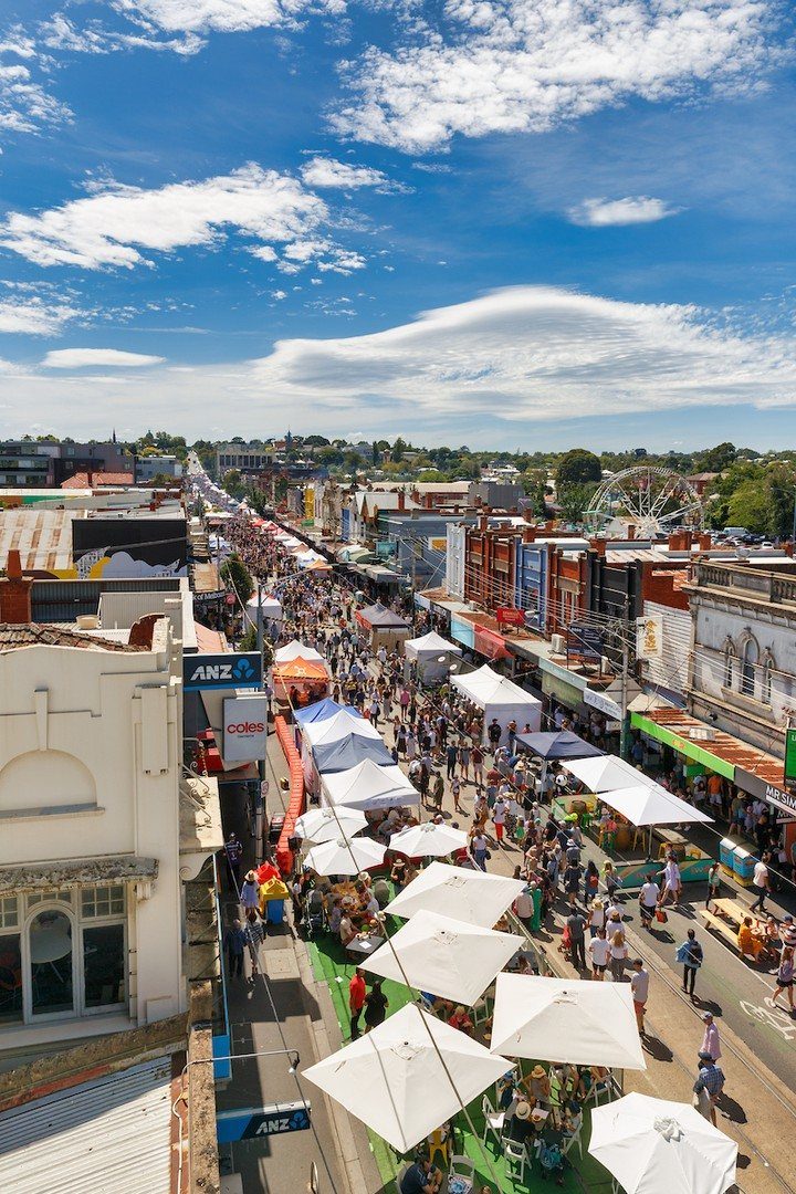 Crowded city street festival with white tents and many people under a blue sky.