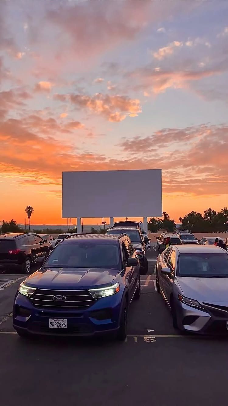 Cars parked in front of a large drive-in movie screen during a colorful sunset.