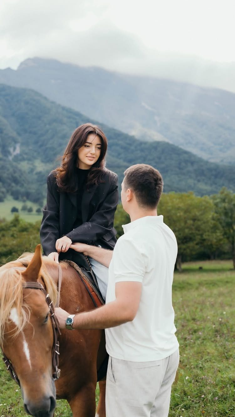 Woman sitting on a brown horse with a man standing beside her outdoors.