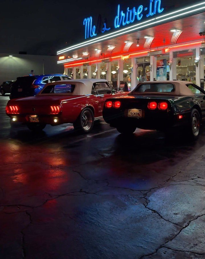 Two vintage convertibles parked before a retro drive-in diner with neon lights at night.