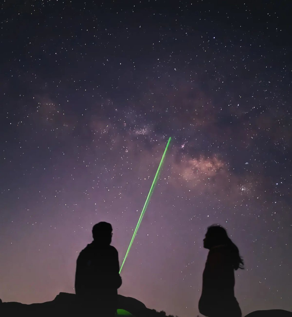 Couple silhouetted against a starry night sky with a green laser pointing upward.