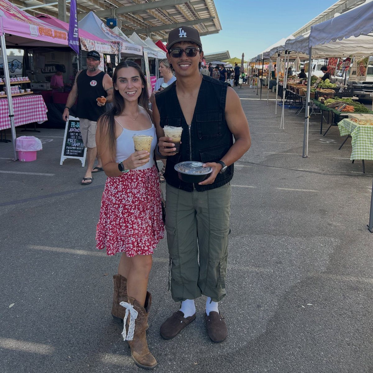 Couple holding drinks and food at an outdoor farmers market with white tents.