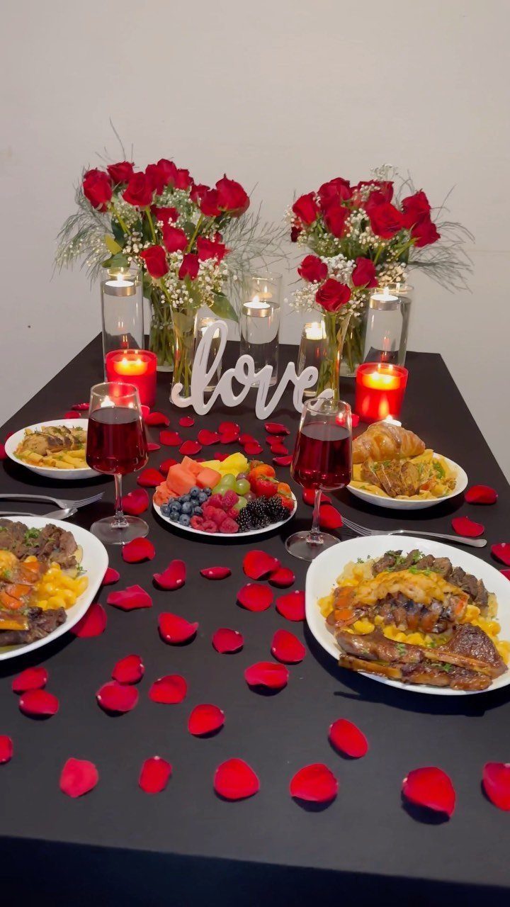 Romantic dinner table with red roses, candles, wine, and plates of food.