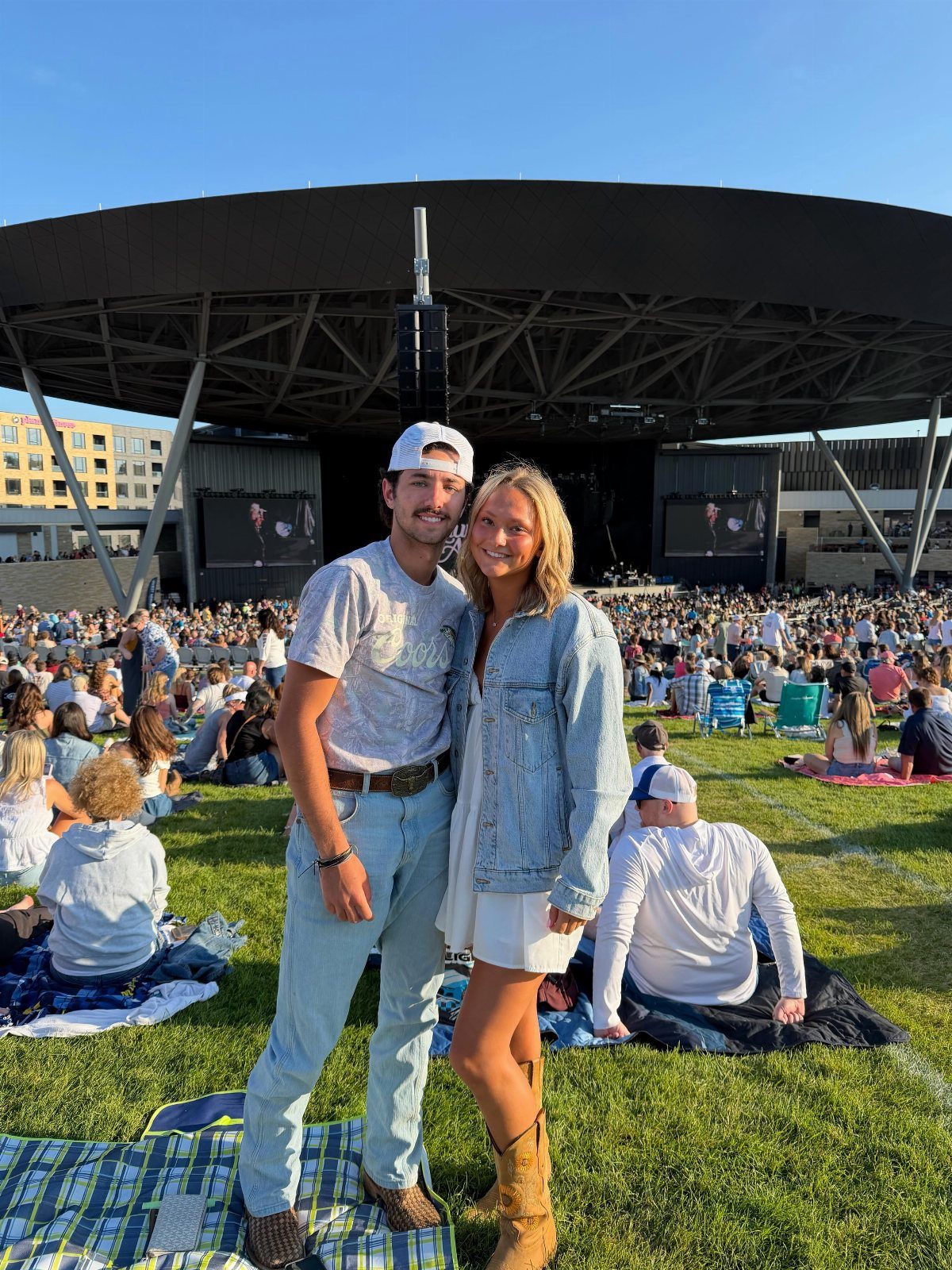 Young couple posing on a grassy lawn in front of a large outdoor concert stage.