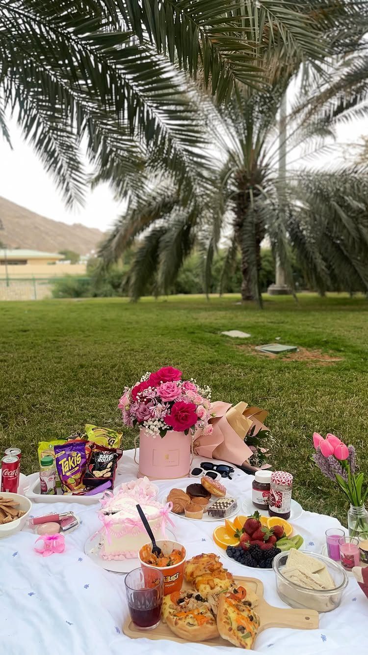 Picnic spread with cake, fruit, and pink roses on a white blanket in a park.