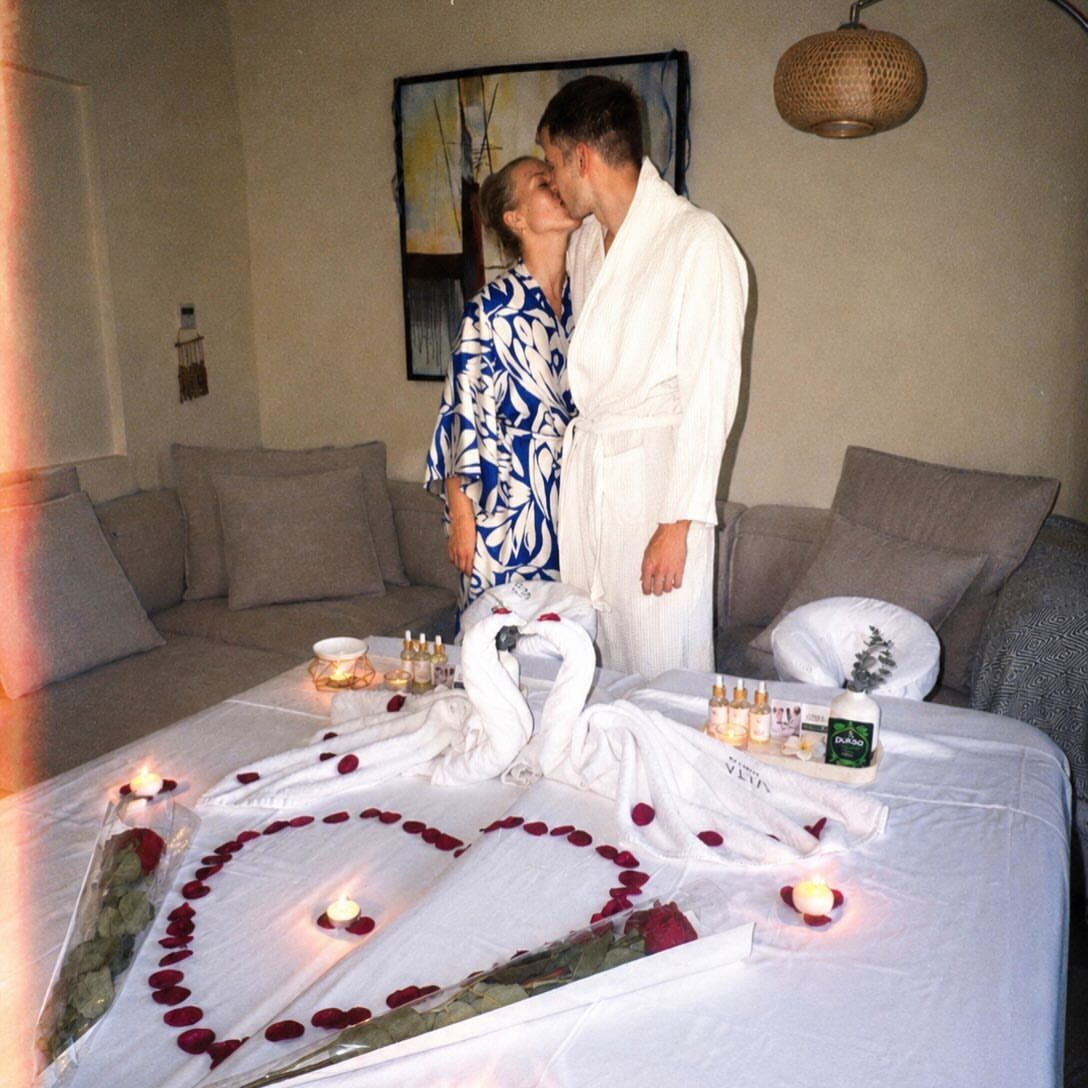 Couple in robes kissing beside a bed decorated with towel swans and rose petals.