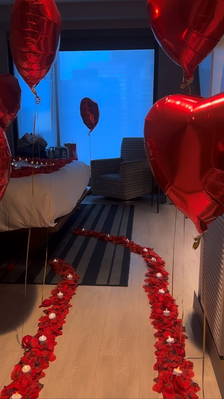 Romantic bedroom with red heart balloons, rose petal paths, and flickering candles.