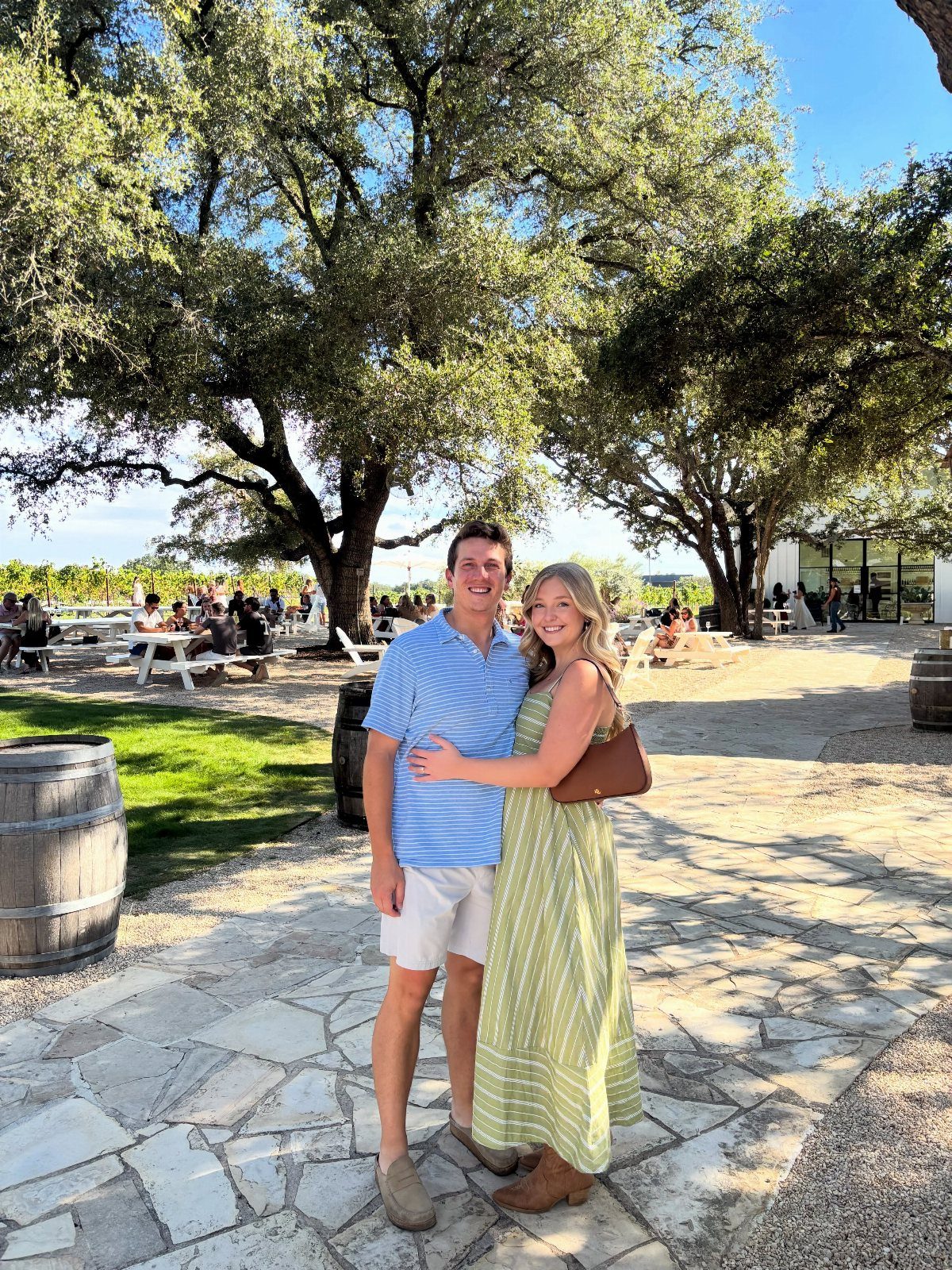 Smiling couple posing on a stone patio with wine barrels and vineyards behind them.