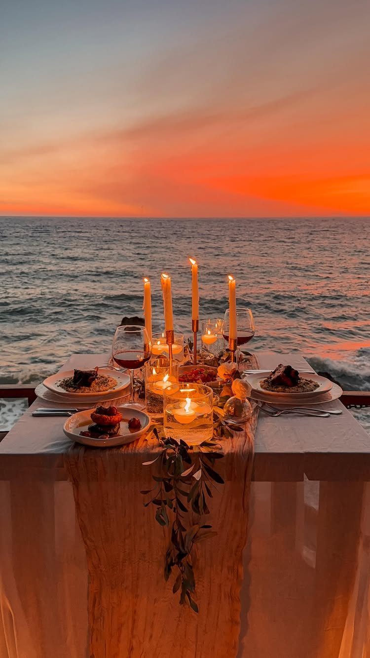 Romantic dinner table with candles and wine overlooking a vibrant ocean sunset.