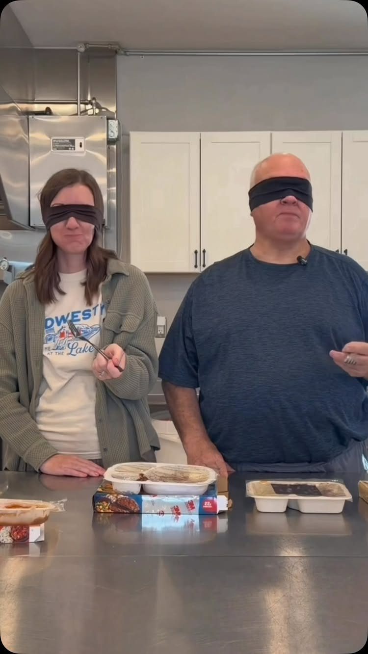 Blindfolded man and woman standing in a kitchen with food containers for tasting.