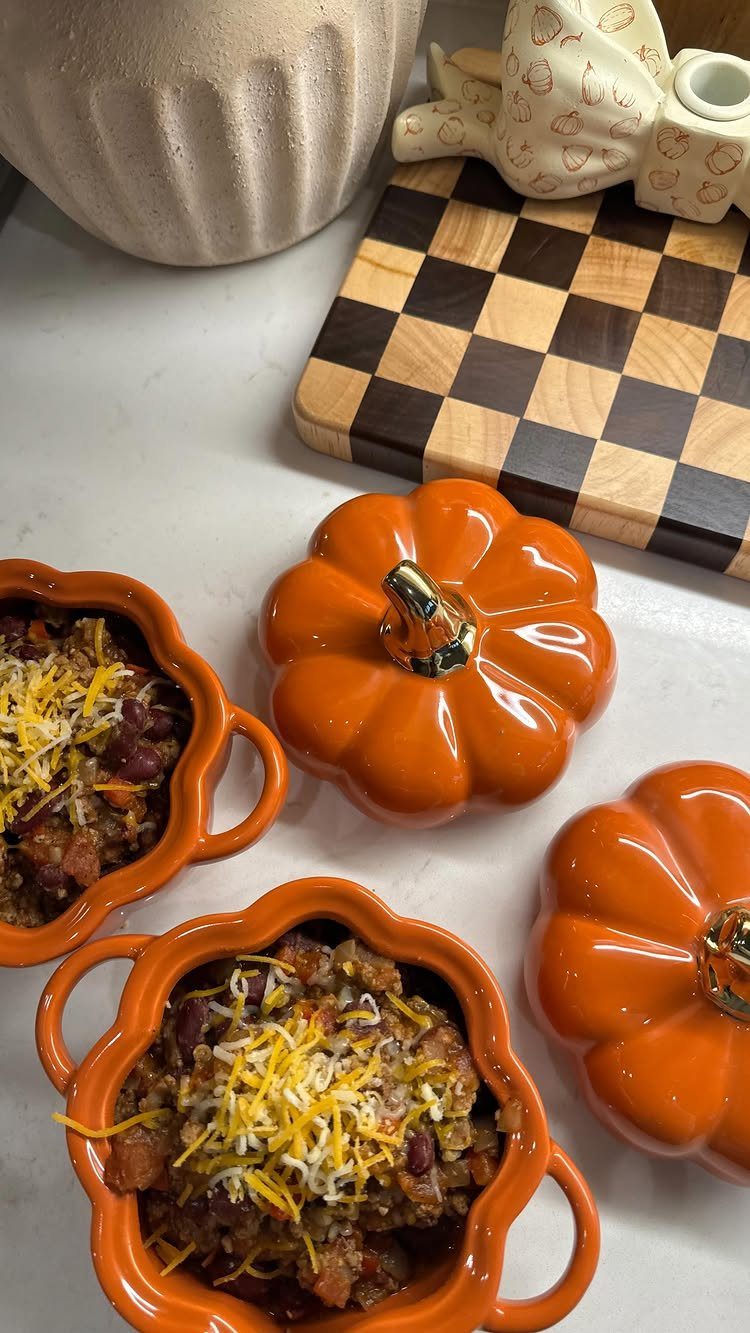 Pumpkin-shaped ceramic bowls filled with cheesy savory food next to a wooden checkerboard.