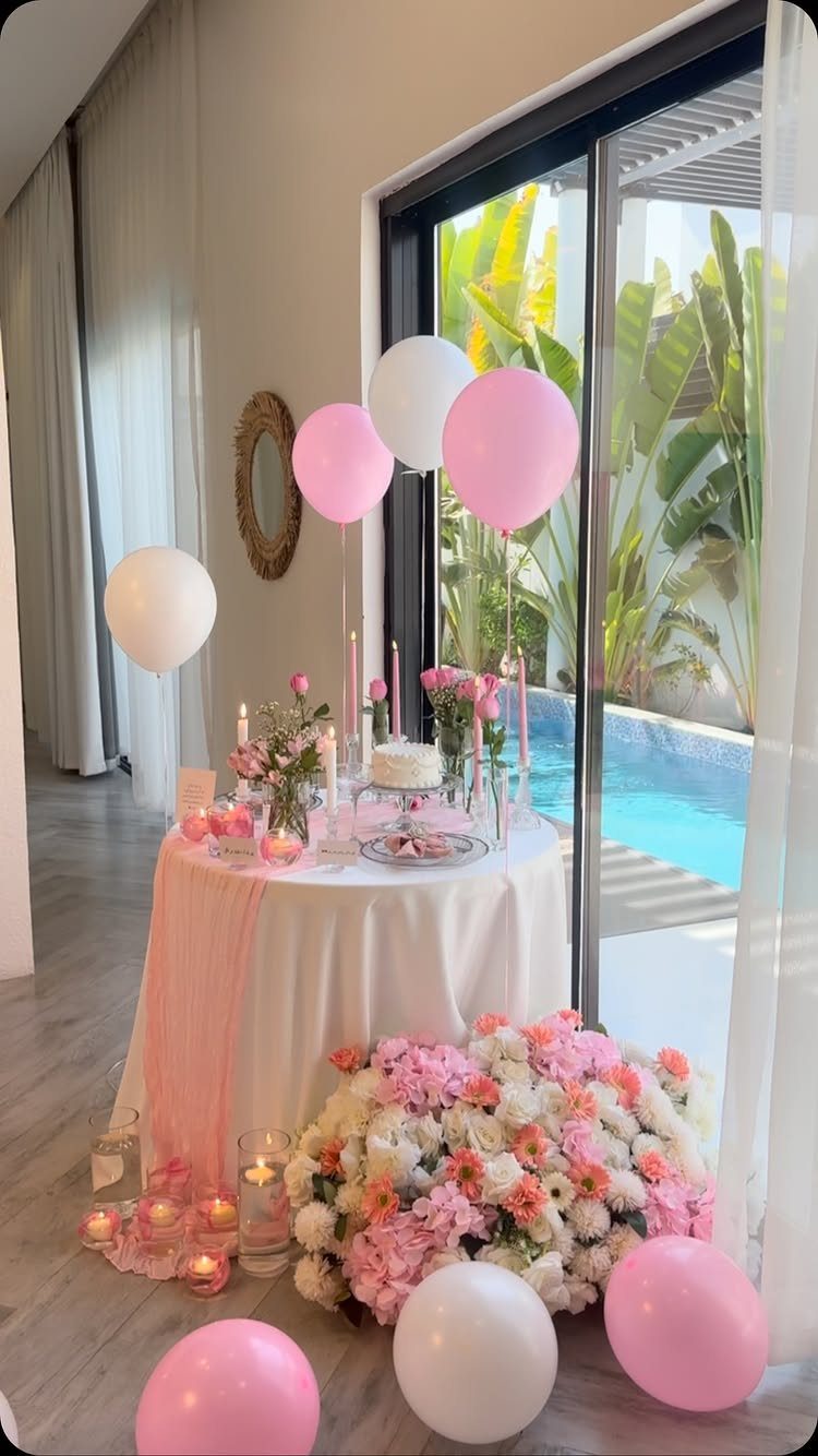 Romantic table setup with pink balloons, flowers, and cake by a pool window.