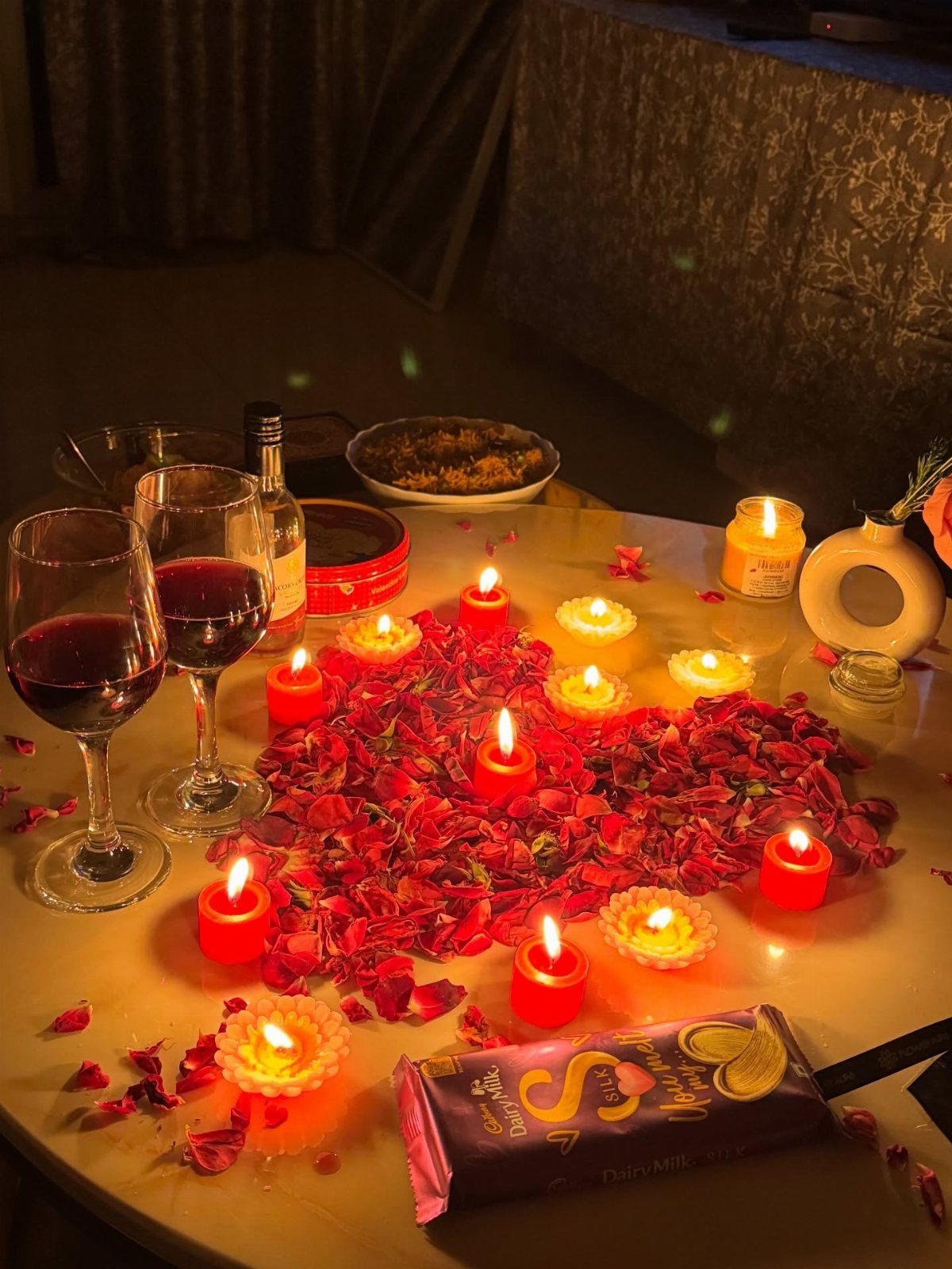 Romantic table setup with red wine, heart-shaped rose petals, and glowing candles.
