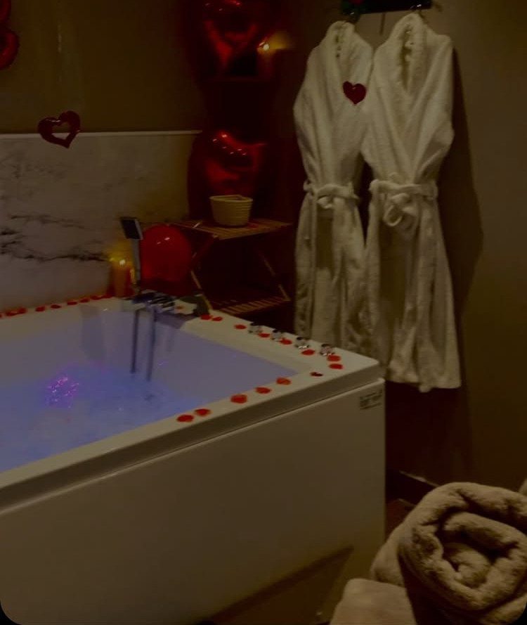 White bathtub with rose petals and two white bathrobes hanging on the wall.