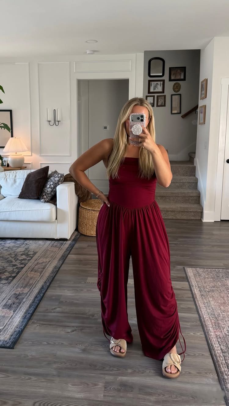 Woman wearing a sleeveless burgundy wide-leg jumpsuit and beige sandals in a living room.