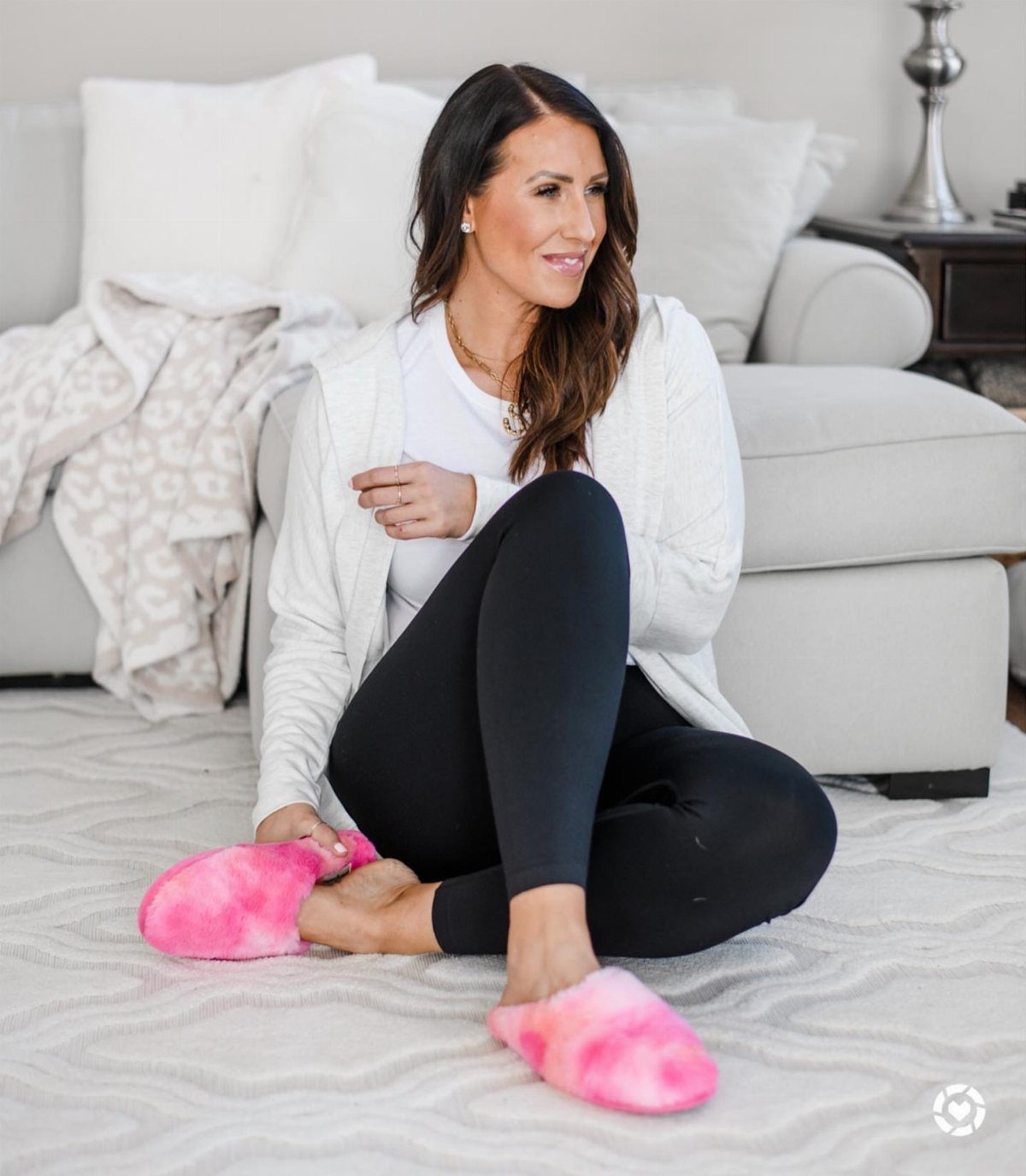 Woman sitting on a white rug wearing pink fluffy slippers and black leggings.