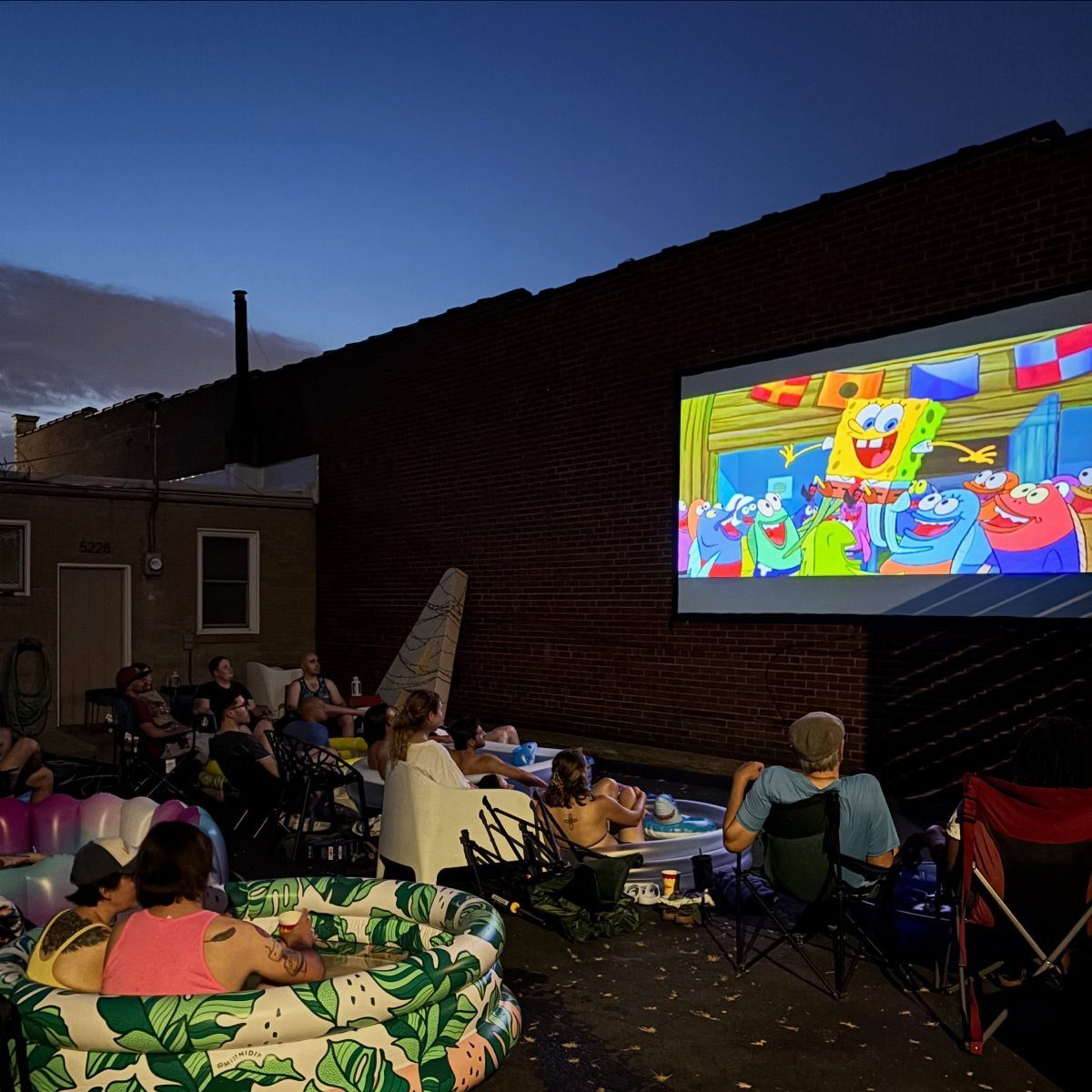 People watching a Spongebob movie on a large outdoor screen against a brick wall.