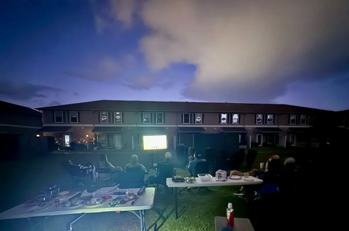 People gathered on a lawn watching a movie on an outdoor screen at dusk.