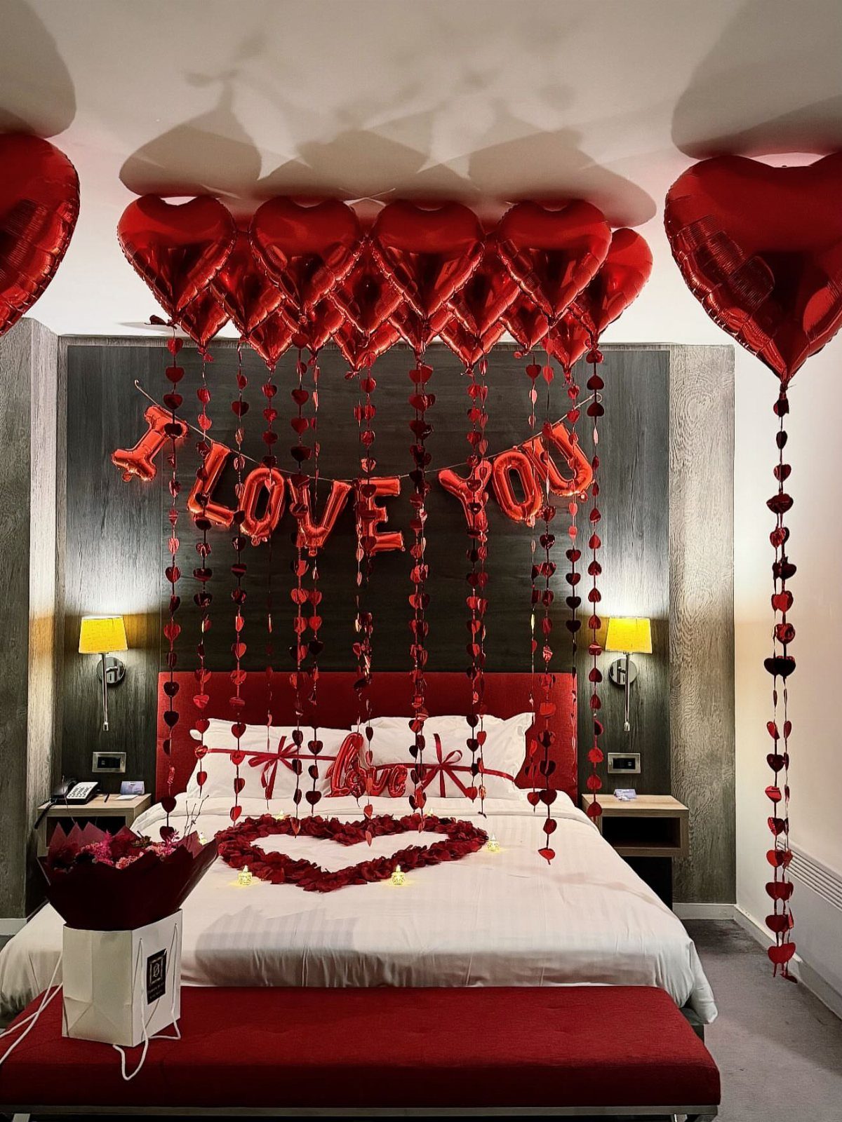 Red heart-shaped balloons floating above a bed decorated with rose petals and banners.