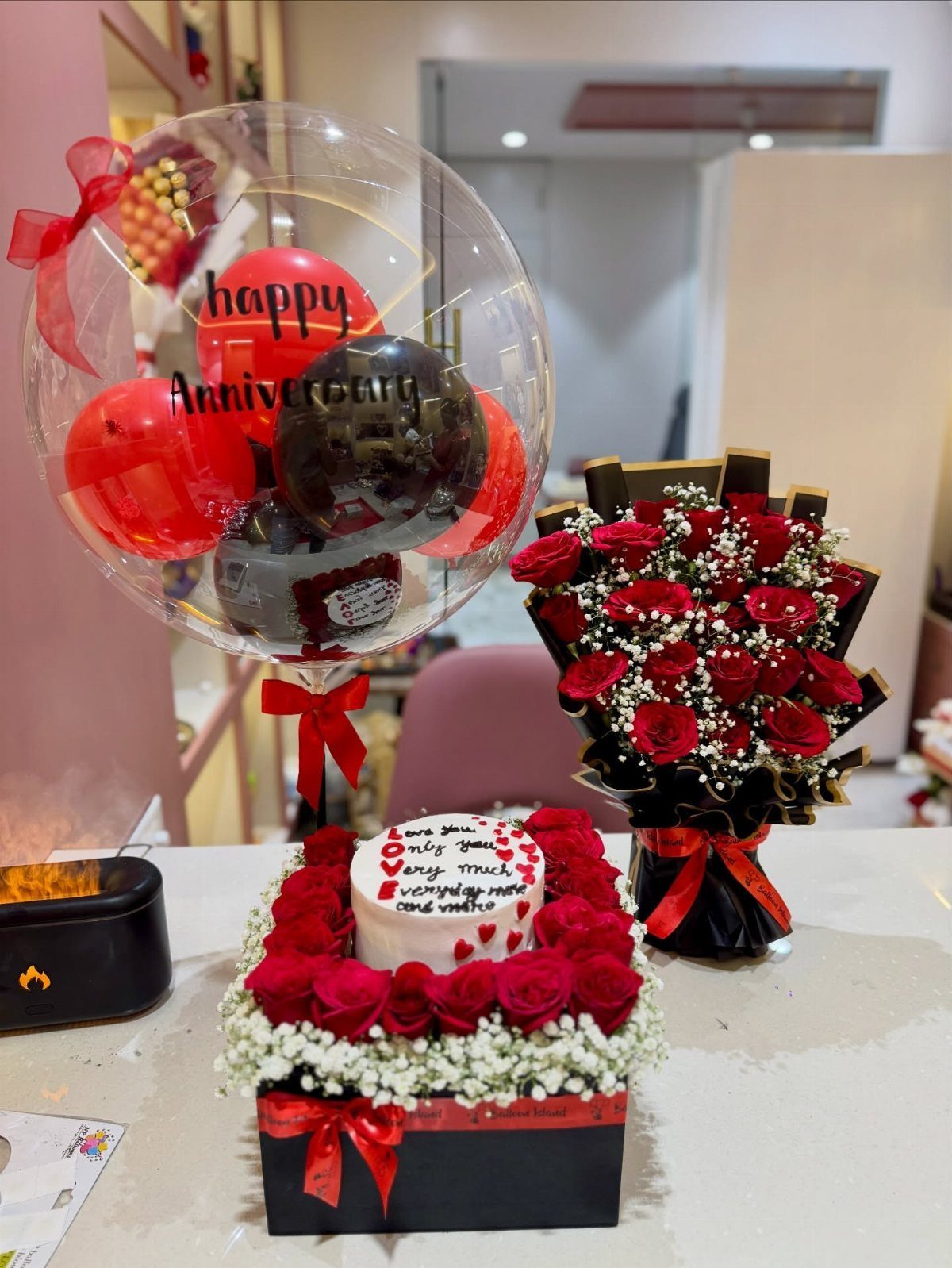 Anniversary gift set with a bubble balloon, red rose bouquet, and heart-shaped cake.