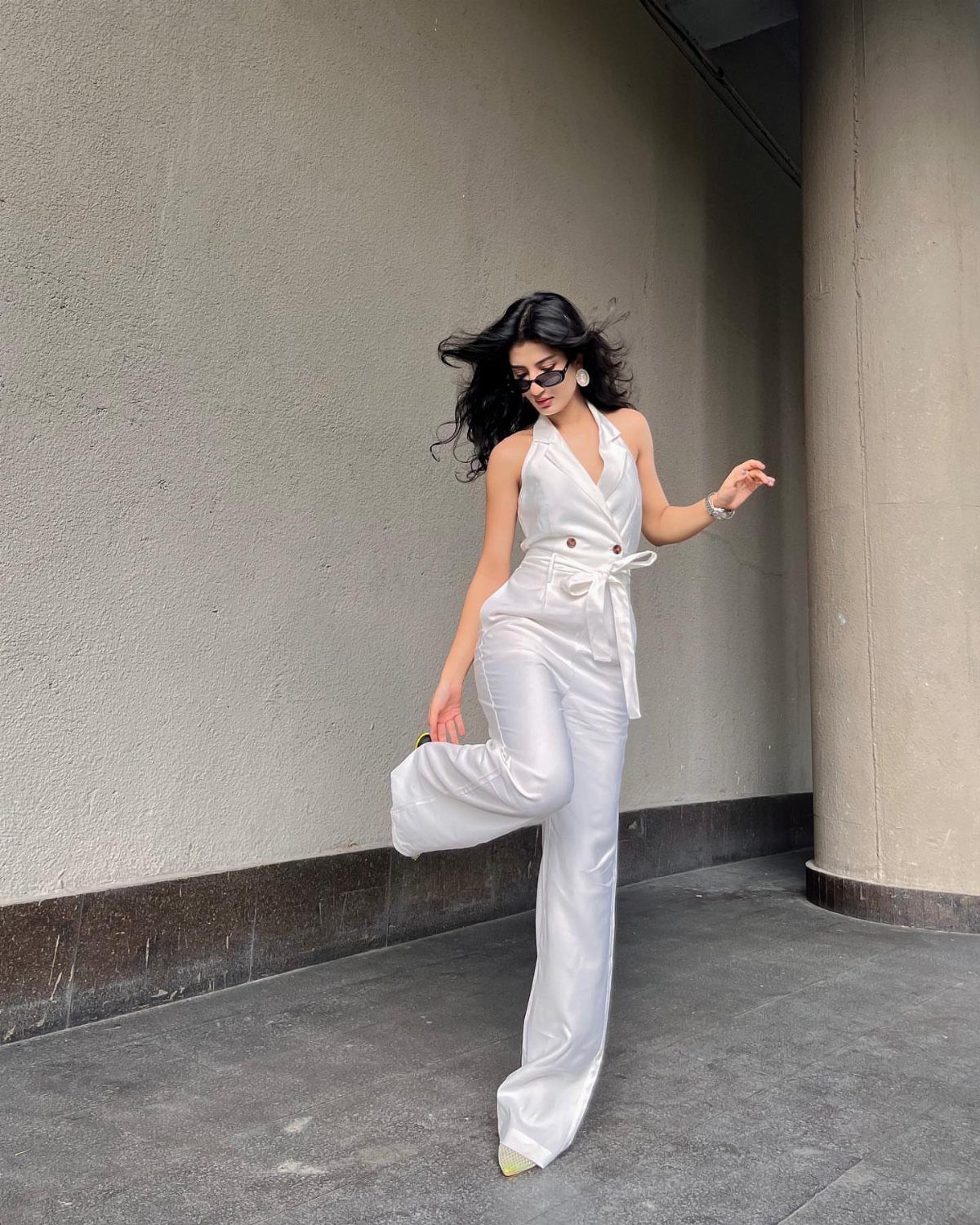 Woman in a white satin sleeveless jumpsuit posing against a grey concrete wall.