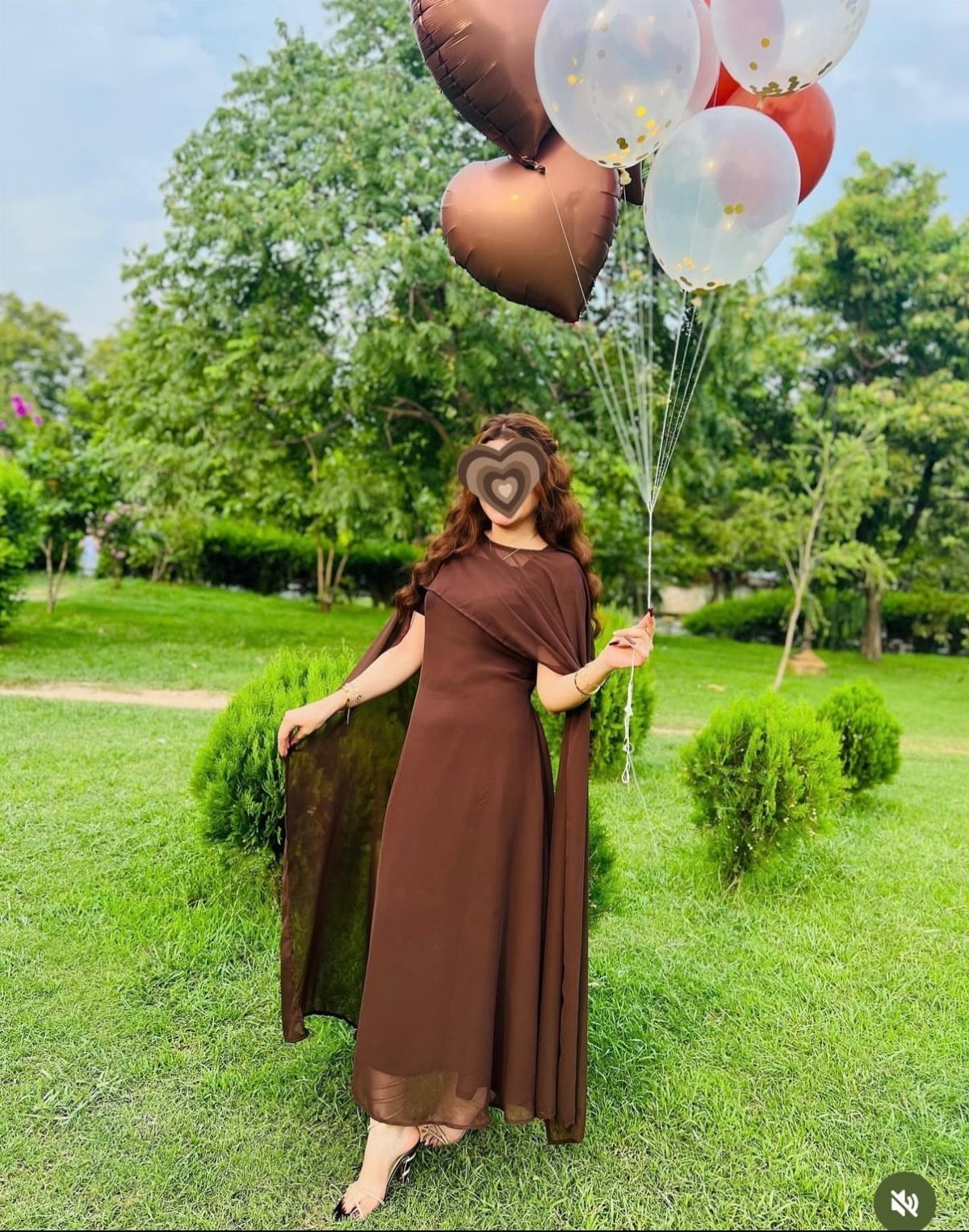 Woman in a long brown flowy dress holding heart-shaped balloons in a park