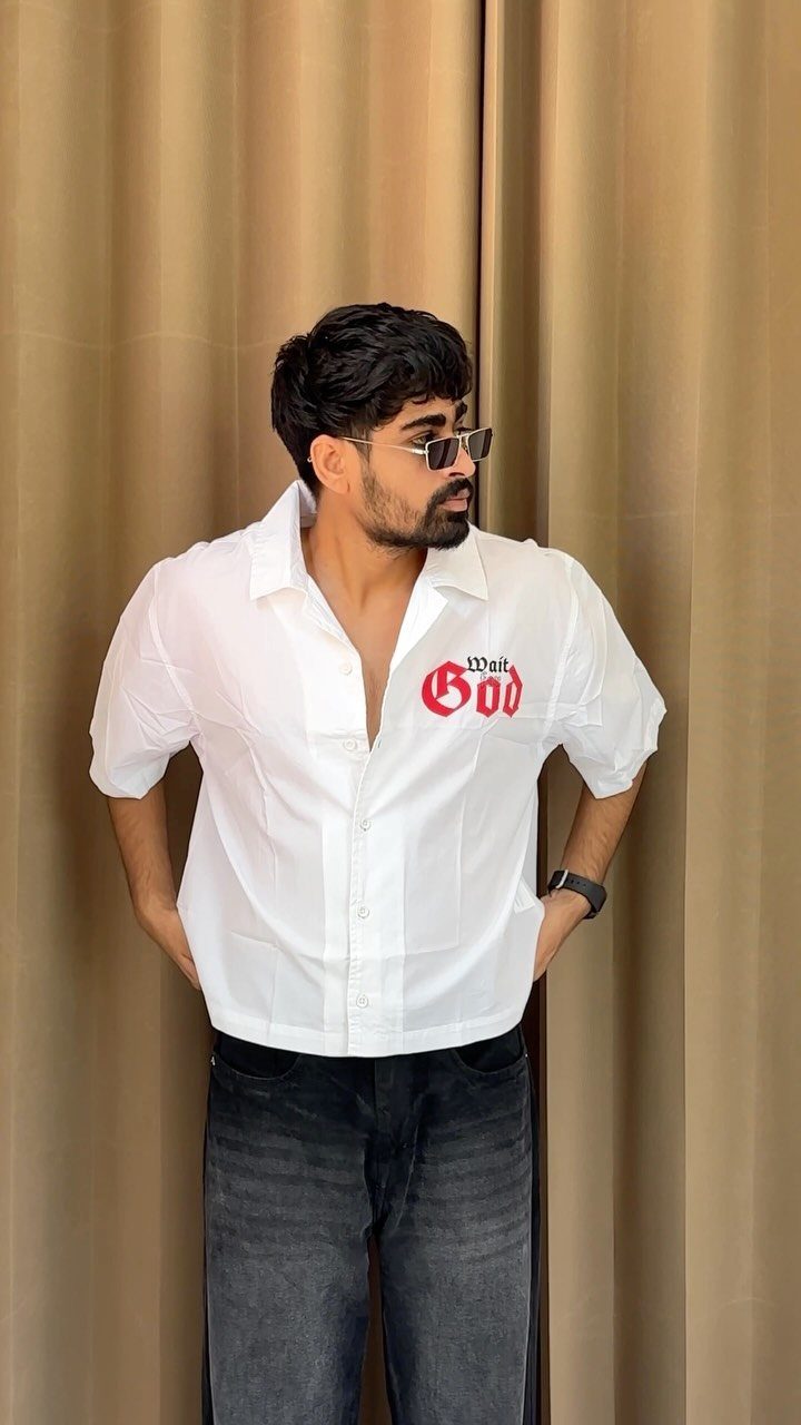 Man wearing a white graphic button-down shirt and dark grey jeans against gold curtains.