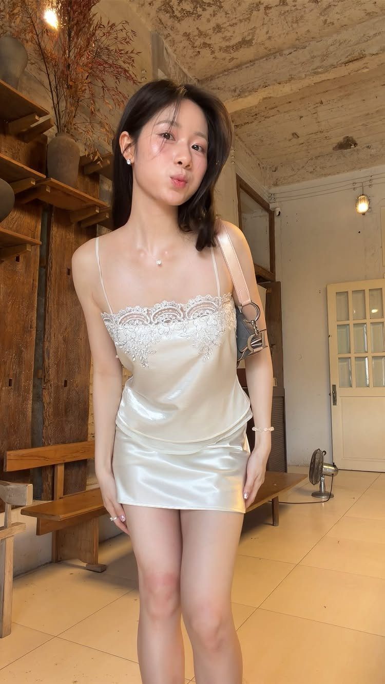 Woman wearing a short white satin slip dress with lace trim in a room.