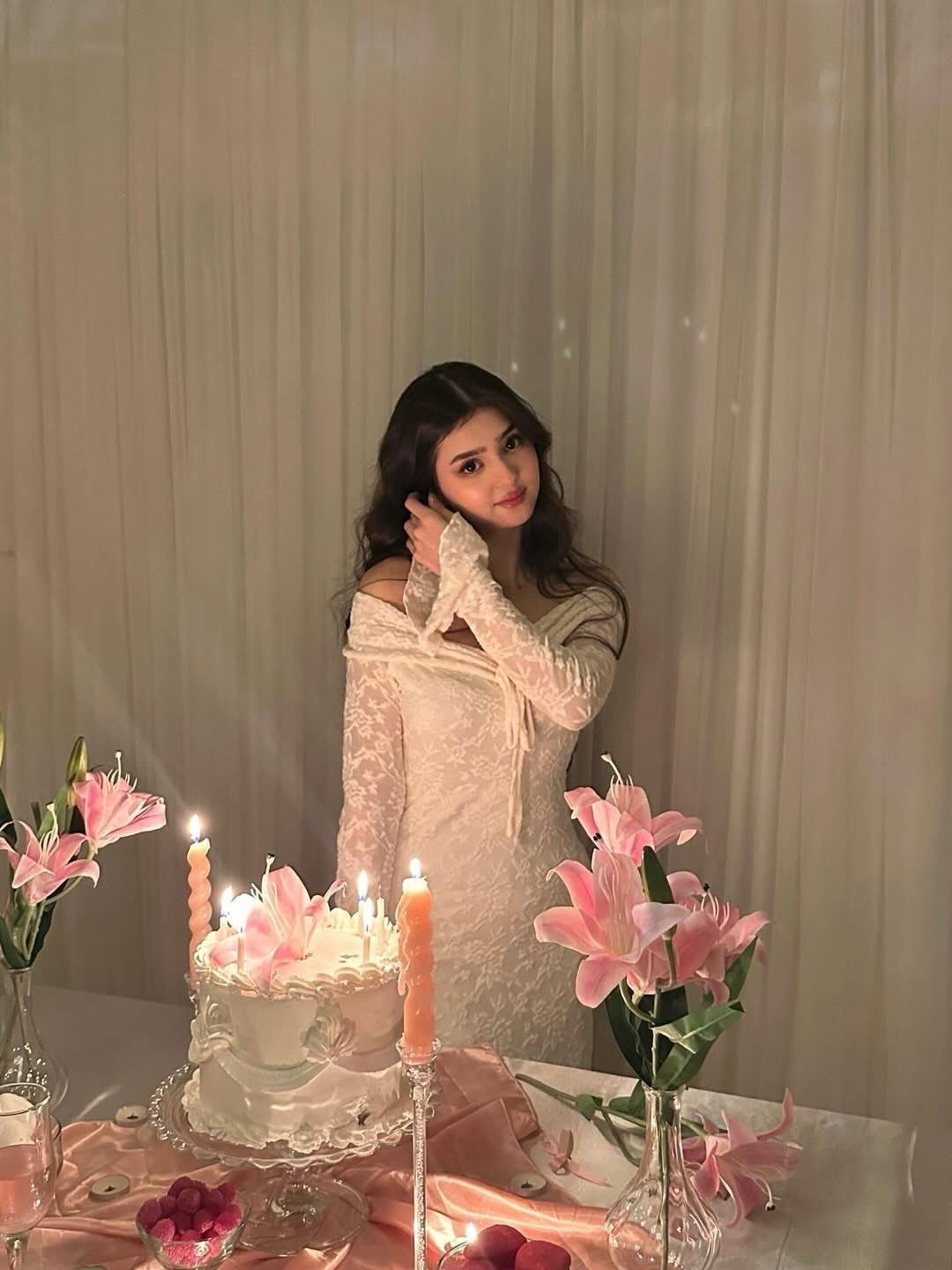 Woman in white lace dress standing behind a birthday cake and pink lilies.