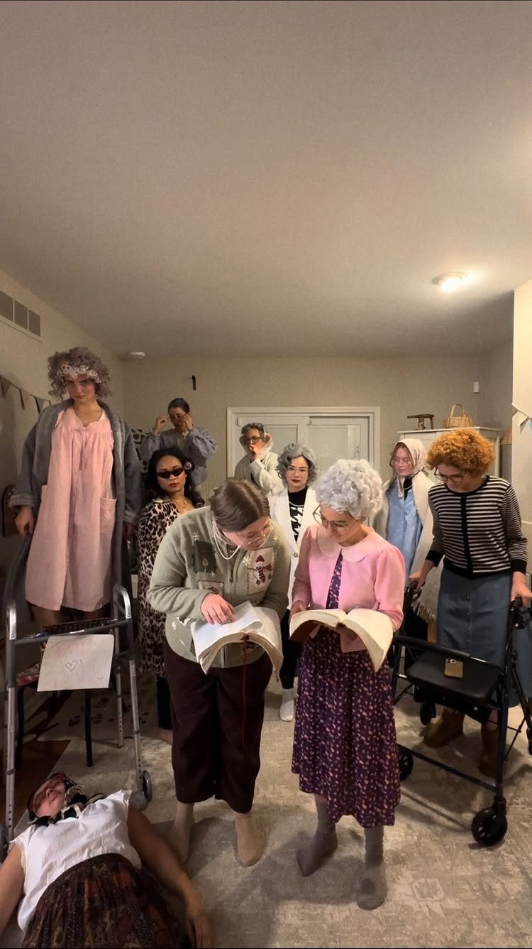 Adults dressed as elderly people with grey wigs and walkers in a living room.