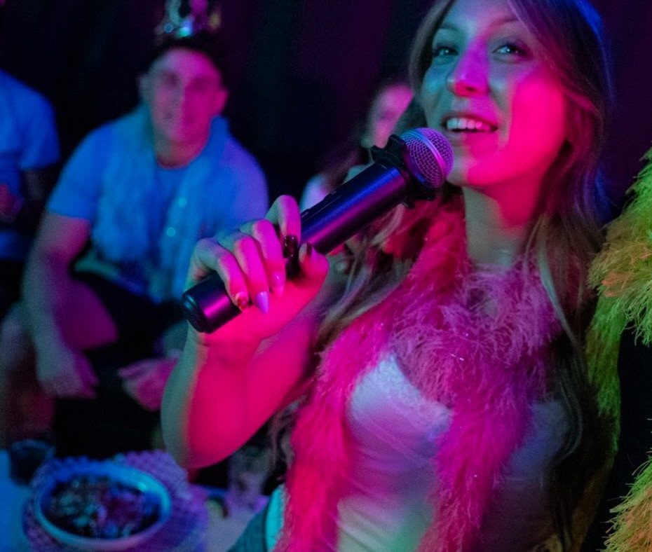 Woman singing into a microphone wearing a pink feather boa at a party