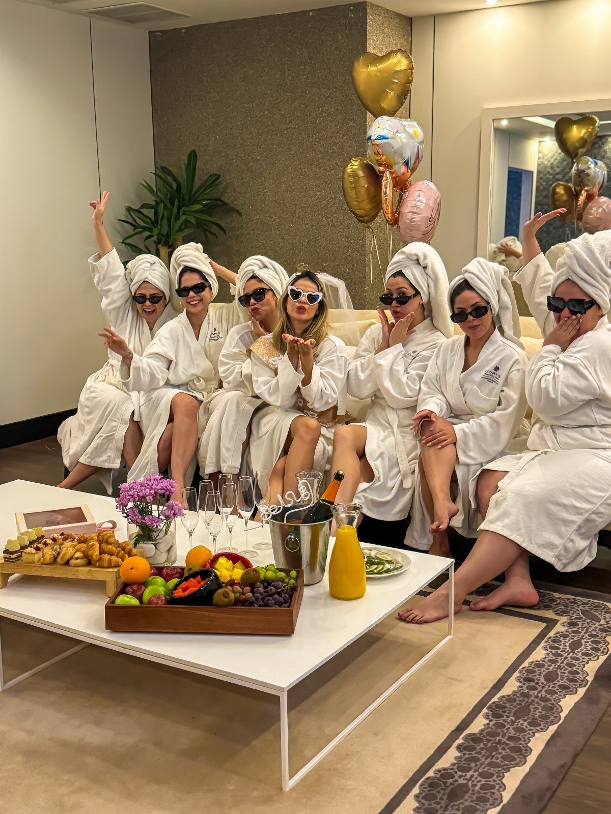 Women in white robes and hair towels relaxing with champagne and fruit.