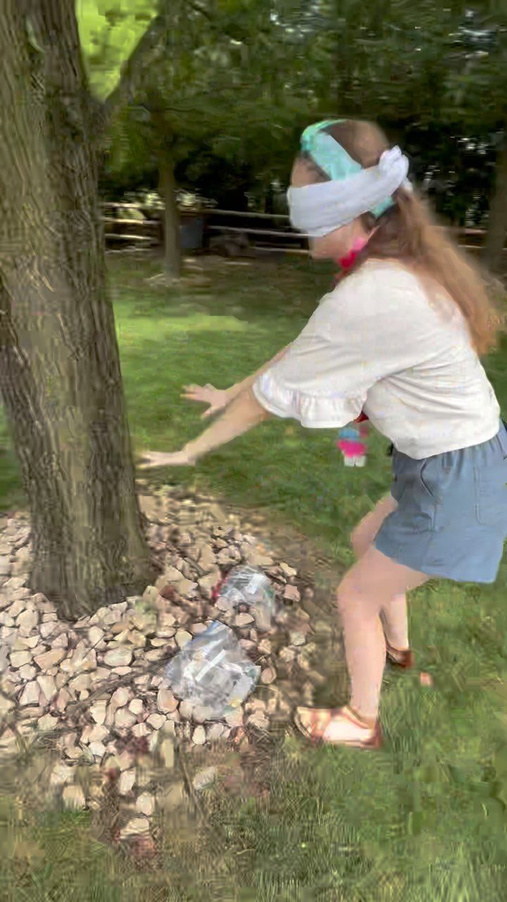 Blindfolded girl reaching forward while standing in a grassy yard next to a tree