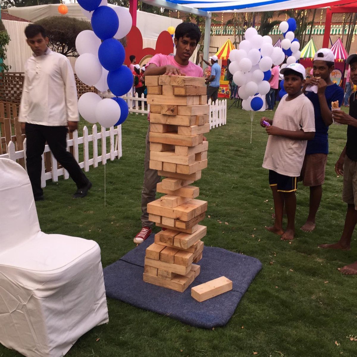 Young man playing giant Jenga on a grassy lawn with balloons in background