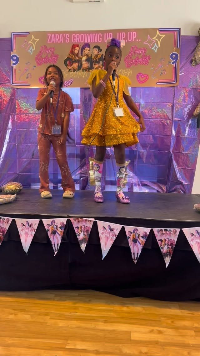 Two young girls singing into microphones on a stage at a birthday party.
