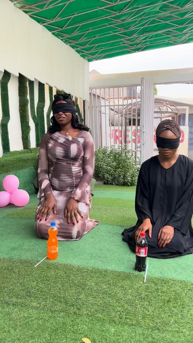 Two blindfolded women kneeling on green grass with drink bottles in front of them