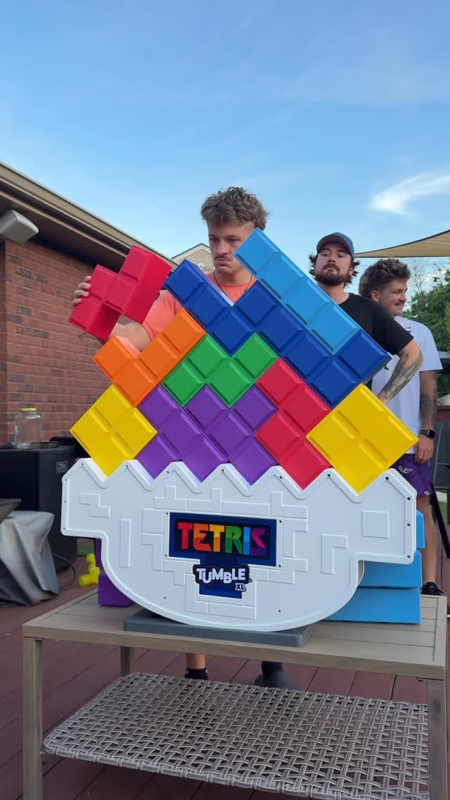 Three men playing a giant Tetris Tumble block game on an outdoor table.