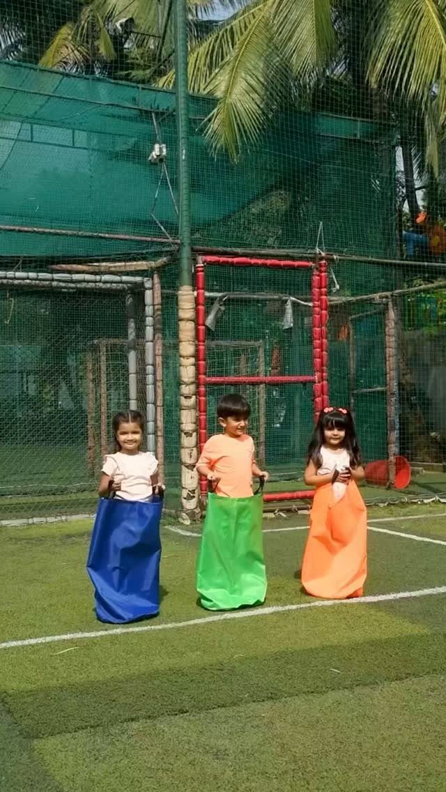 Three children standing in blue, green, and orange sacks on a green field.