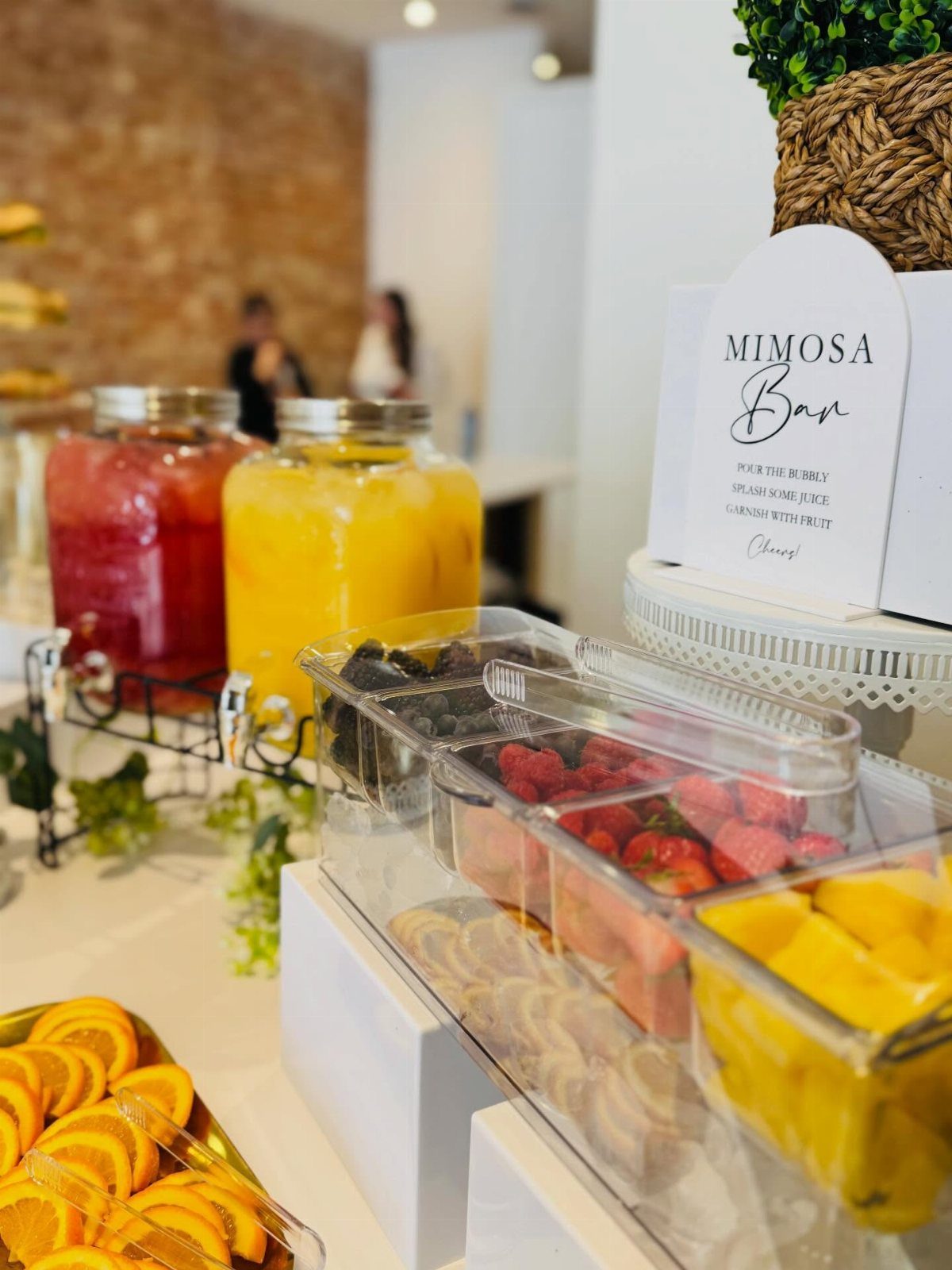 Mimosa bar with juice dispensers and fresh fruit toppings on a white table.