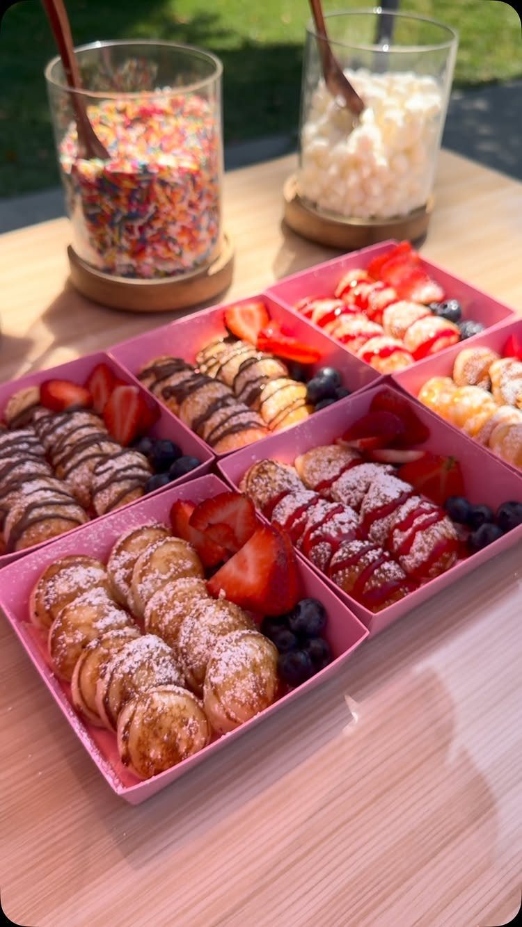 Mini pancakes with fruit and toppings served in small pink square boxes.
