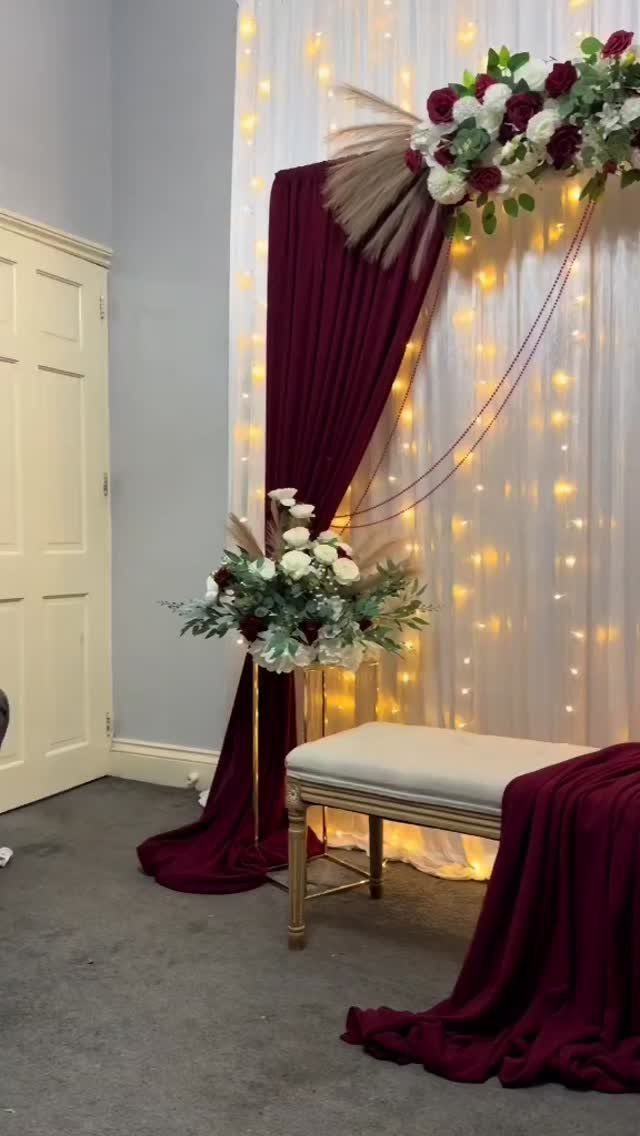 Photo backdrop with white fairy lights, burgundy drapery, floral arrangements, and a bench.