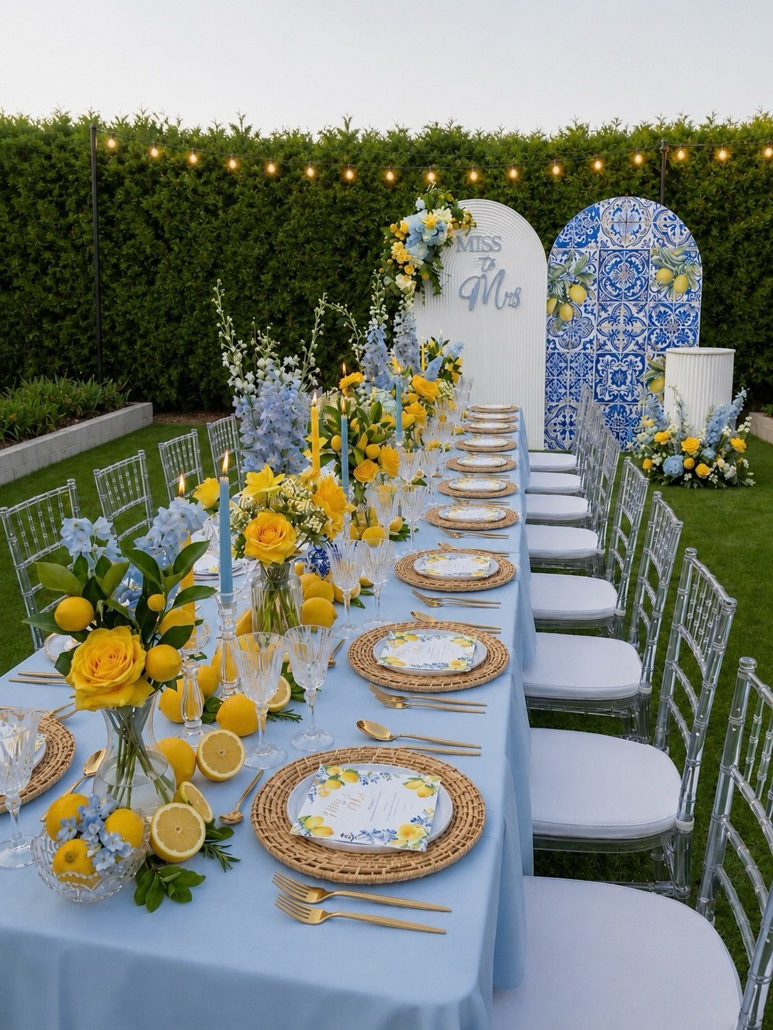 Long outdoor table set with blue linens, yellow roses, and fresh lemons.