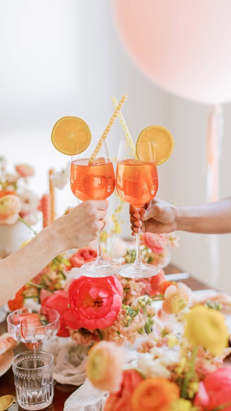 Two orange cocktails with lemon slices clinking over a table of bright flowers.