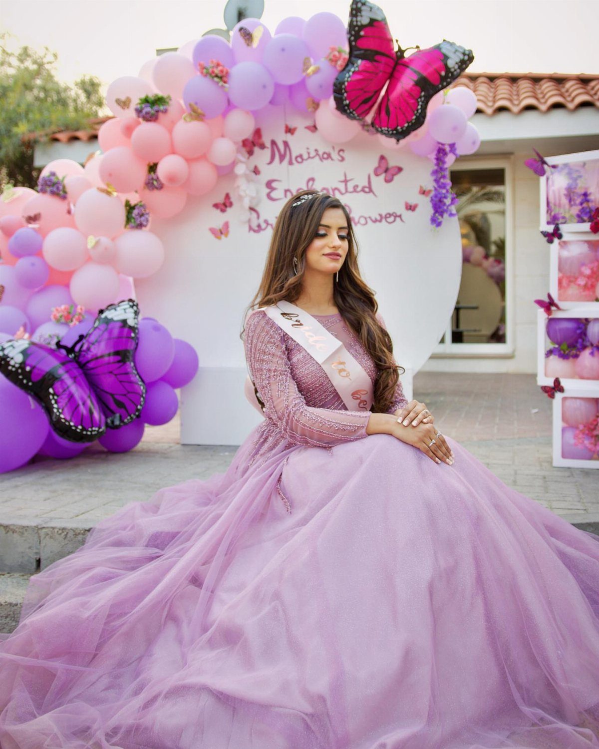 Woman in purple gown sitting before a whimsical backdrop with balloons and butterflies.