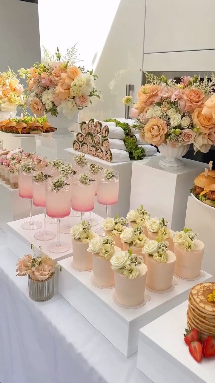 Pink cocktails and mini cakes on white tiered stands with peach rose bouquets.