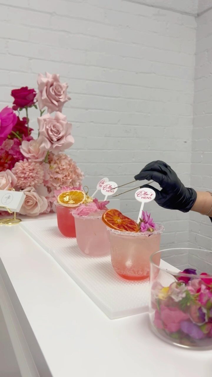 Pink cocktails with citrus garnishes being prepared next to a pink rose bouquet.