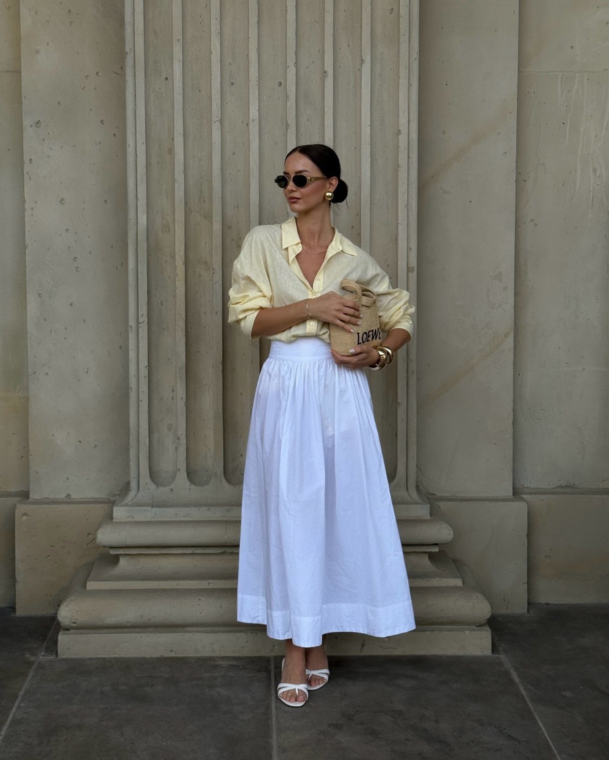 Woman in yellow button-down shirt and long white pleated skirt standing by stone column.