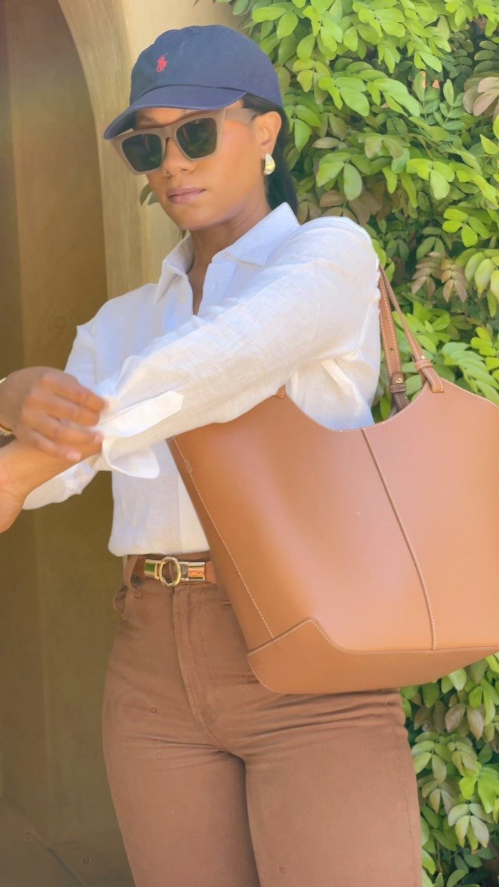 Woman in white button-down shirt and brown trousers carrying a tan leather tote bag.