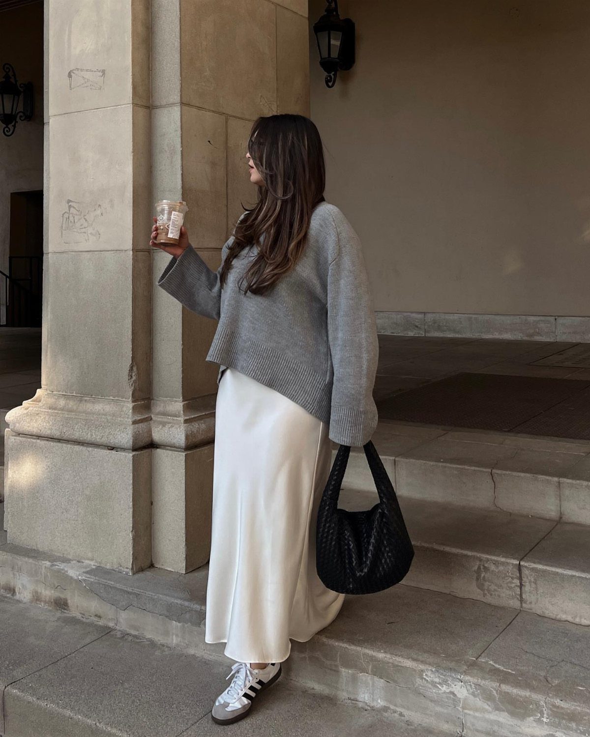 Woman in grey knit sweater and white satin skirt standing on stone steps
